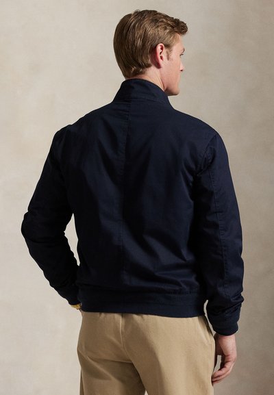 Bărbat purtând o geacă bleumarin și pantaloni bej, stând cu spatele către cameră pe un fundal simplu.