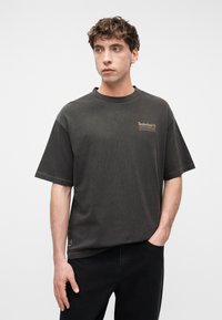 Νέος άνδρας με κατσαρά μαλλιά, φορώντας μια άνετη σκούρα γκρι μπλούζα Timberland και μαύρο παντελόνι, στέκεται μπροστά σε λευκό απλό φόντο.