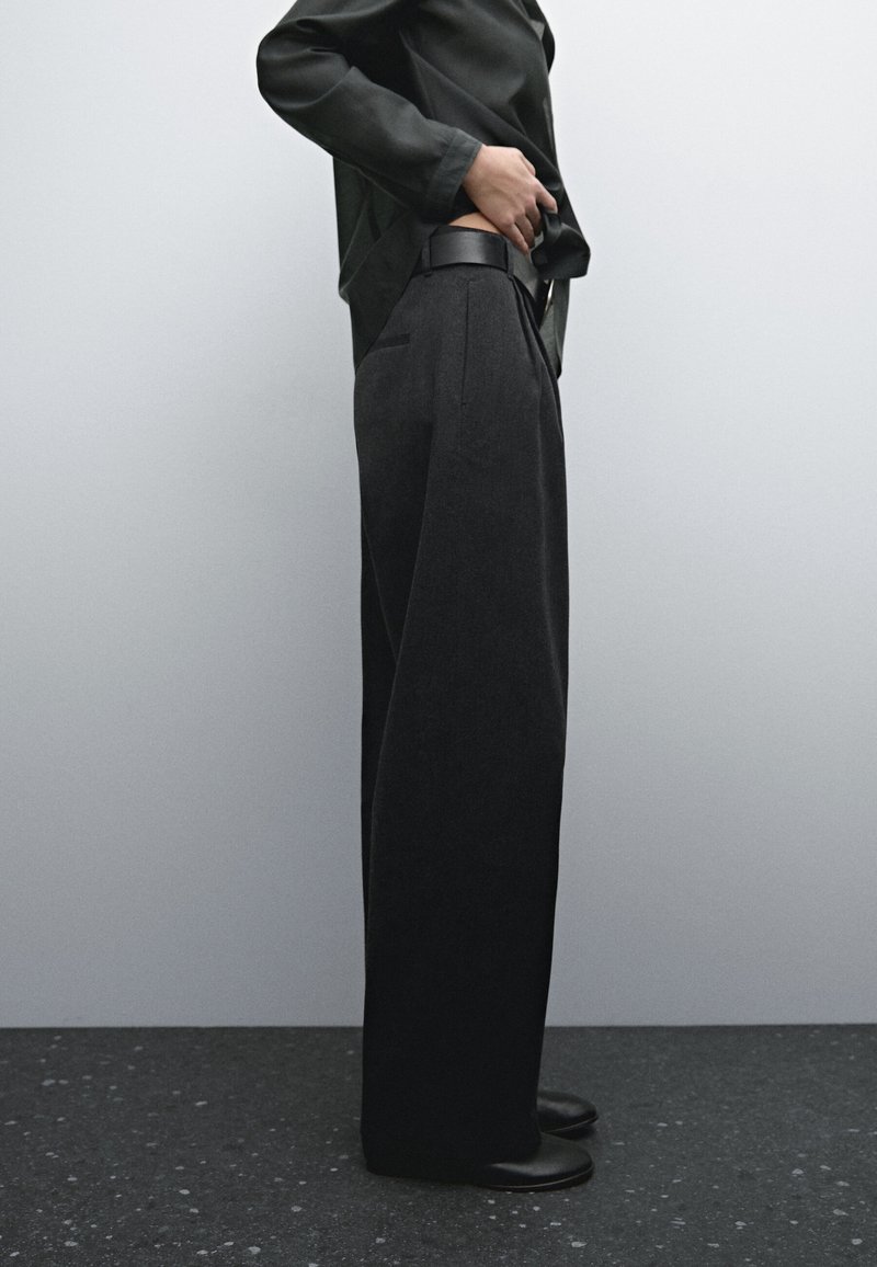 Personne portant un pantalon noir à jambes larges, une ceinture noire, un haut noir et des chaussures noires, debout de profil sur un fond clair uni.