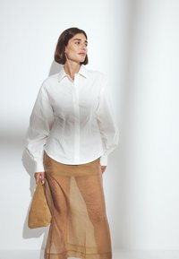 Witte blouse met lange, opgezette mouwen, gecombineerd met een doorschijnende, lichtbruine midi-rok. De outfit heeft een zachte, textuurrijke afwerking.