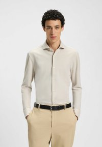 Lichtbeige blouse met lange mouwen, een puntkraag en knoopsluiting, gecombineerd met textuurbeige broeken en een zwarte riem.