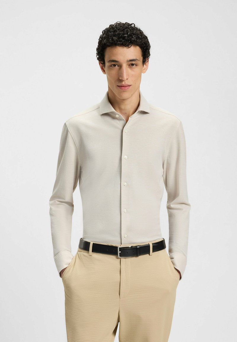 Lichtbeige blouse met lange mouwen, een puntkraag en knoopsluiting, gecombineerd met textuurbeige broeken en een zwarte riem.