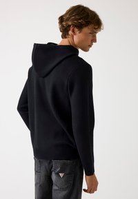 Felpa nera con cappuccio realizzata in un tessuto strutturato, design aderente e tasca a marsupio, abbinata a jeans grigi con un logo applicato sul retro.