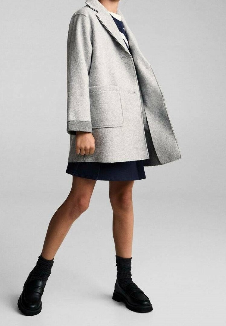 Personne portant un manteau en laine gris clair sur une jupe bleu foncé, des chaussettes noires et des mocassins noirs, debout devant un fond uni gris clair.