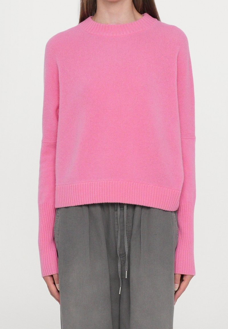 Femme portant un pull en maille côtelée rose vif à manches longues, associé à un pantalon gris ample avec cordon de serrage, debout devant un fond uni.