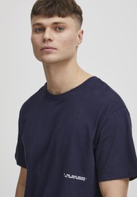 Marineblå bomulds T-shirt med korte ærmer. Har en rund halsudskæring og en lille hvid tekstgrafik nær bunden til venstre.