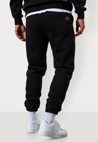 Schwarze Sweathosen aus weichem Stoff mit elastischem Bund und Bündchen, mit einem kleinen Logodetail auf der Rückentasche. Kombiniert mit weißen Turnschuhen.