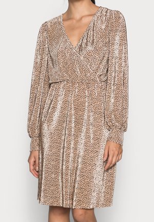 Vrouw draagt een beige jurk tot op de knie met lange mouwen, zwarte polka dots, een overslag met V-hals en gerimpelde manchetten.