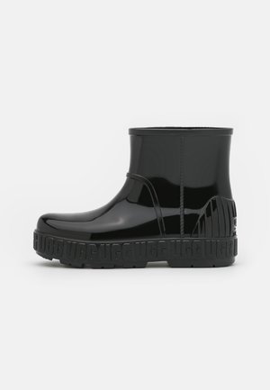 Botte de pluie noire brillante pour cheville avec semelle épaisse embossée du logo "UGG" en répétition et panneau de talon texturé, vue latérale sur fond blanc.
