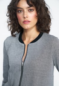 Kobieta z kręconymi brązowymi włosami i piegami, ubrana w szary zamek błyskawiczny z wzorem w jodełkę i czarnym kołnierzem na białym tle.