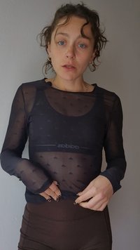 Γυναίκα με σγουρά μαλλιά που φοράει διαφανές μαύρο μακρυμάνικο τοπ πάνω από αθλητικό σουτιέν Adidas και σκουρόχρωμο κολάν, διορθώνοντας το πουκάμισό της.