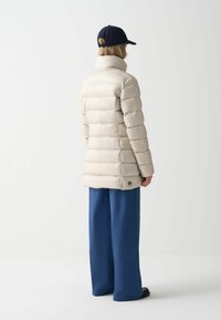 Chaqueta acolchada beige con cuello alto, diseño acolchado y dobladillo con puños, combinada con pantalones anchos azules. La vista trasera resalta la textura y el ajuste.