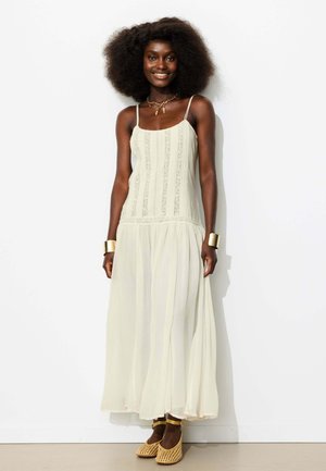 Femme avec une coiffure afro naturelle portant une robe maxi crème sans manches, des bracelets dorés, des colliers superposés et des chaussures tissées à bride cheville, debout et souriante.
