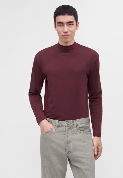 Jeune homme aux cheveux courts noirs portant un haut bordeaux à manches longues et col montant, ainsi qu'un jean gris clair, debout avec une main dans la poche.