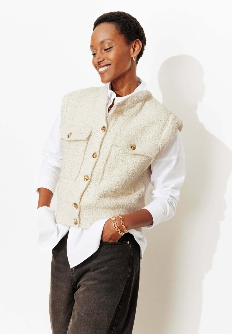 Gilet tricoté couleur crème avec deux poches avant, boutons dorés et bords côtelés, porté par-dessus une chemise blanche à boutons, associé à un pantalon foncé.