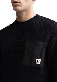 Pull en tricot noir texturé avec une poche lisse contrastante portant une étiquette. Encolure ronde et poignets côtelés complètent le design.