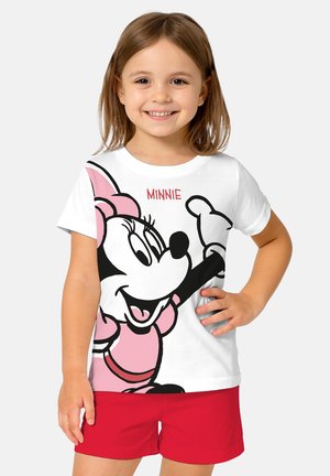 Lächendes Mädchen mit braunen Haaren trägt ein weißes T-Shirt mit Minnie Mouse in pinkfarbenem Kleid und rote Shorts, steht mit einer Hand in der Hüfte.