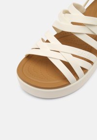 Bílé sandály s popruhy, které mají měkkou hnědou stélku a silnou bílou podrážku. Design zahrnuje hladkou texturu a vyhýbá se jakýmkoli viditelným kovovým prvkům.