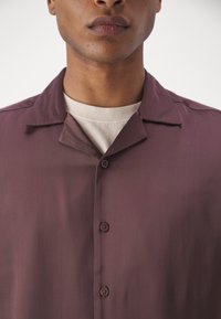 Chemise bordeaux à manches courtes en tissu lisse, dotée d'un col camp, d'une patte de boutonnage et d'une texture douce. Portée par-dessus un haut de couleur claire.