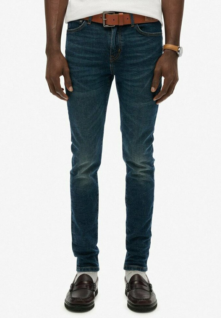 Hombre vistiendo jeans oscuros ajustados, cinturón de cuero marrón, reloj de cuero marrón, calcetines blancos y mocasines marrones oscuros, de pie contra un fondo blanco.