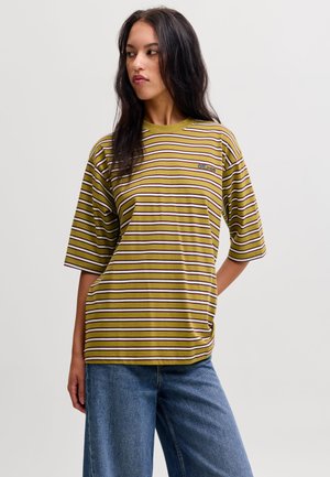 Femme aux longs cheveux foncés portant un t-shirt ample rayé vert, blanc et marron et un jean bleu, debout devant un fond uni.