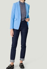 Ljusblå blazer över en mönstrad mörkblå blus. Mörka jeans i denim med uppvikta ärmar och glänsande svarta loafers med metallaccenter.