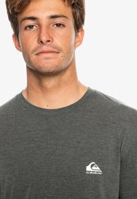 Sivá dlhá mikina s dlhým rukávom vyrobená z mäkkej textílie, s malým bielym logom Quiksilver na ľavej strane hrude a s klasickým okrúhlym výstrihom.