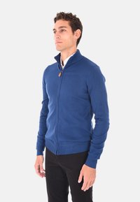 Maglione blu con zip, colletto alto, polsini e orlo a coste. Texture liscia, materiale leggero, con un tirazip in pelle.