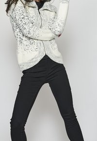 Stickad cardigan i vit och grå mönster, med dragkedja framtill och ribbade ärmslut. Matchas med svarta skinny jeans med subtila dekorationer.