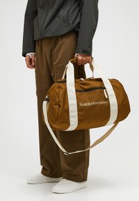 Bolso de lona marrón con correas y cremallera blancas. Hecho de tela, presenta un logo en el lateral. Persona que lleva pantalones marrones lo sostiene.