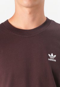 Hnedé bavlnené tričko s okrúhlym výstrihom a bielym logom Adidas s tromi pruhmi nad ním, hladká textúra a štandardný strih.