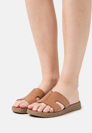 Voeten in tan leren instapsandalen met uitsparingen en gevoerde bruine zolen op een witte achtergrond.