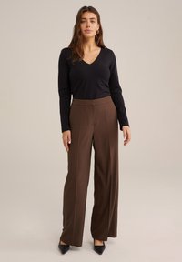 Schwarzes Langarmoberteil mit V-Ausschnitt, kombiniert mit hoch taillierten, braunen weit geschnittenen Hosen. Spitz zulaufende schwarze Absätze runden das Outfit ab.