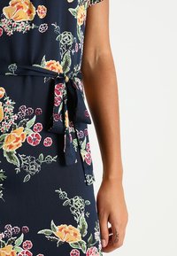 Robe en tissu navy avec imprimé floral, présentant des manches courtes et un détail de taille nouée. Le design comprend diverses fleurs colorées et feuillage.