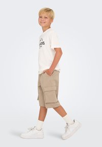 T-shirt blanc avec imprimé graphique noir, shorts cargo khaki avec poches latérales, baskets blanches et chaussettes blanches. Tenue décontractée pour enfants.