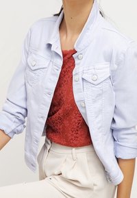 Lichtblauwe denimjacket met twee borstzakken, witte knopen en een ronde kraag, gedragen over een rood kanten top. Soepel van textuur.