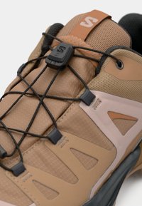 Μπεζ πεζοπορικό παπούτσι με υφασμάτινο πάνω μέρος, μαύρα ελαστικά κορδόνια και λαστιχένια σόλα. Διαθέτει εμφανές σύστημα κορδονιών και διακοσμητικά από δίχτυ.