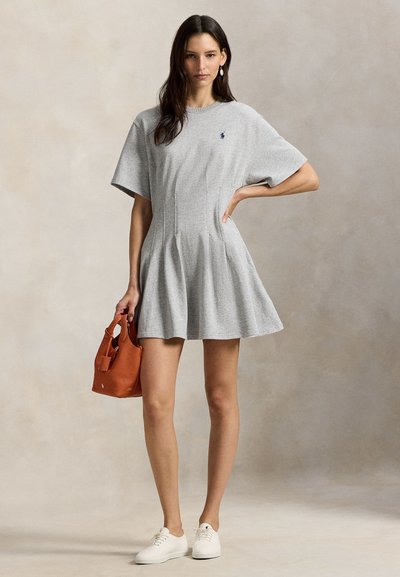 Γυναίκα που φοράει γκρι φουσκωτό φόρεμα με κοντά μανίκια, λευκά αθλητικά παπούτσια, κρατώντας μια μικρή καφέ δερμάτινη τσάντα, στέκεται μπροστά σε ουδέτερο φόντο.