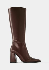 Wysoki brązowy skórzany but za kolano z zaostrzonym noskiem i szerokim, blokowym obcasem, pokazany z boku na jednolitym białym tle.