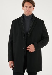 Abrigo de lana negro con un diseño estructurado, que presenta dos botones, bolsillos laterales y un forro interior negro con un patrón. Lleva puesto sobre un cuello de tortuga oscuro.