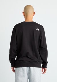 Svart hettegenser med ribbestrikket krage og mansjetter, med en liten hvit logo på baksiden. Sammen med lysegrå joggebukser.