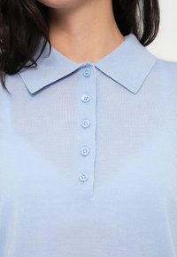 Ανοιχτό μπλε πόλο μπλούζα με απαλή υφή, που διαθέτει κλασικό γιακά και σειρά από έξι κουμπιά σε τόνους που ταιριάζουν, κατά μήκος του μπροστινού μέρους.
