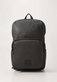 Sac à dos en nylon noir avec une surface texturée, doté d'une poche frontale zippée et d'une poignée rembourrée sur le dessus. Patch avec le logo à l'avant.