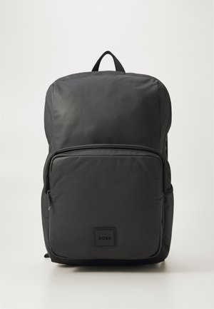 Sac à dos en nylon noir avec une surface texturée, doté d'une poche frontale zippée et d'une poignée rembourrée sur le dessus. Patch avec le logo à l'avant.