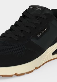 Черен кец Skechers с текстурирани детайли, черни връзки, бяла междинна подметка и кафява подметка с показано името на марката отстрани и на езика.