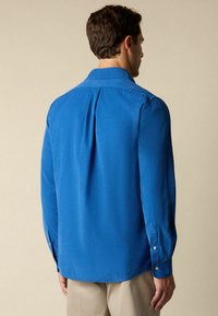 Man die met zijn rug naar de camera staat, gekleed in een lichtblauw overhemd met lange mouwen en beige broek, tegen een effen beige achtergrond.