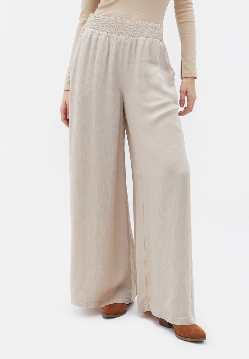 Väljer en vidbenad, beige byxa i mjukt tyg med texturerad yta; har resårmidja och en lös passform i kombination med bruna skor.
