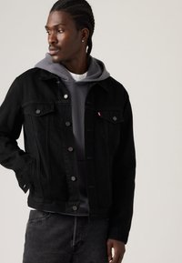 Čierna rifľová bunda s zapínaním na gombíky, vreckami na hrudi a bočnými vreckami na ruky. Nosená cez sivú mikinu s kapucňou. Neformálny strih.