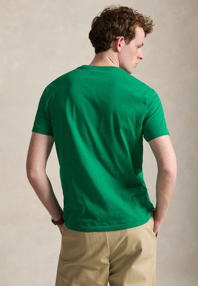 Mand med kort krøllet hår iført en grøn kortærmet skjorte og beige bukser, stående med hænderne i lommerne, vendt væk.