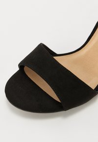 Sandalia de ante negra con diseño de punta abierta, que presenta una tira ancha sobre el pie y una plantilla de color claro. Textura suave, con detalles mínimos.